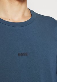 Modré bavlněné tričko s kulatým výstřihem a krátkými rukávy, s vyvýšeným černým logem na přední straně, které zní "BOSS".