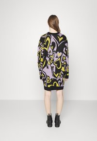 Abito maglione nero con vivaci motivi a cuore in viola e giallo. Tessuto a maglia testurizzato; maniche lunghe e orlo a coste. Indossato con stivali neri alla caviglia.