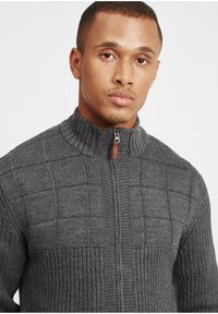 Grå strikket cardigan med høy krage, med glidelås og teksturert sjakkmønster. Stoffet er mykt, med ribbede detaljer.