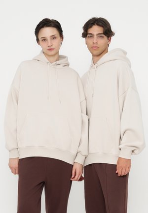 Dos personas de pie una al lado de la otra con sudaderas con capucha beige y pantalones marrones sobre un fondo blanco liso.