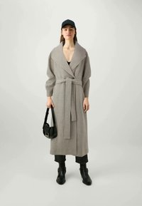 Cappotto grigio lungo con cintura, colletto ampio e maniche lunghe; indossato sopra un top nero e jeans scuri, abbinato a una borsa nera e stivali.