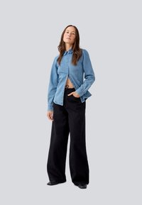 Lys blå denimskjorte med krage og knapper, båret åpen for å avdekke midjen; paret med brede svarte bukser. Glatt tekstur, avslappet design.