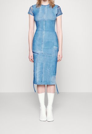 Frau trägt ein eng anliegendes blaues Patchwork-Denimkleid mit kurzen Ärmeln und weiße Stiefel bis zur Mitte der Wade, sie steht vor einem einfarbigen Hintergrund.