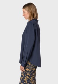 Camicia a maniche lunghe blu navy con colletto, dotata di polsini con bottoni e un taglio rilassato. Abbinata a una gonna a motivi marrone dorato.
