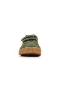 Zapato de ante verde con dos cintas de Velcro ajustables, suela de goma marrón texturizada y panel lateral en amarillo, con diseño de punta redondeada.