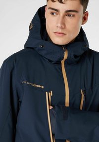 Giacca impermeabile blu navy con accenti dorati, tasche con cerniera e cappuccio perforato. Presenta polsini regolabili e un design aderente.