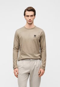 Μπλούζα με μακριά μανίκια σε μπεζ χρώμα, κατασκευασμένη από βαμβάκι, με μαύρο λογότυπο στο στήθος, φορεμένη με ανοιχτό γκρι παντελόνι με ραφές.