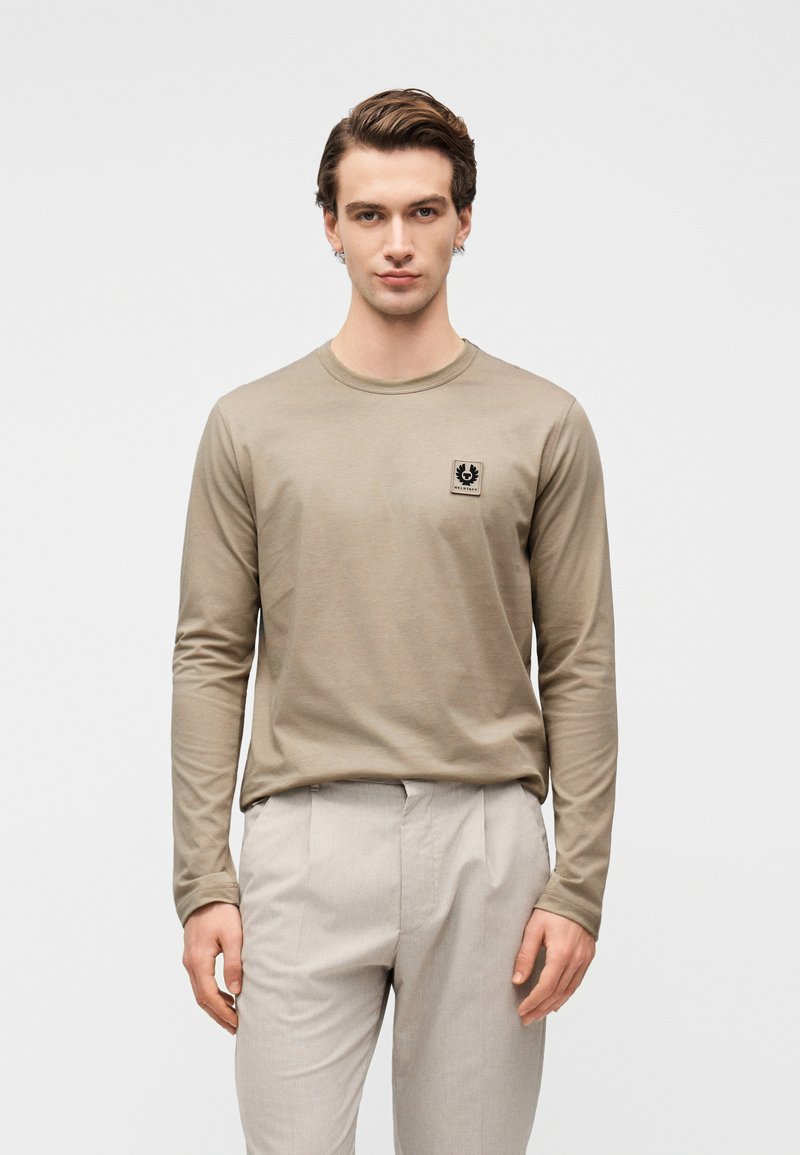 Μπλούζα με μακριά μανίκια σε μπεζ χρώμα, κατασκευασμένη από βαμβάκι, με μαύρο λογότυπο στο στήθος, φορεμένη με ανοιχτό γκρι παντελόνι με ραφές.