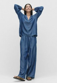 Denim-outfit med en lös, V-ringad topp med långa ärmar och matchande vida byxor. Båda plaggen är i en enfärgad blå nyans med en slät textur.