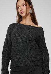 Schwarzer, gerippter Strickpullover mit schulterfreiem Design und langen Ärmeln. Weicher Stoff, cropped Länge. Modell trägt silberne, runde Ohrringe.
