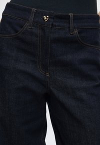 Tmavomodré džínsy z denimu so zlatými srdcovými kovovými prvkami na pásiku, s kontrastným prešívaním a hladkou textúrou.