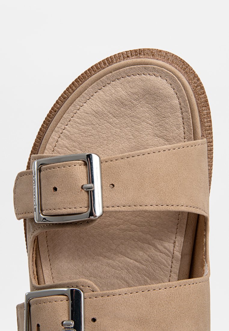 Beige Sandale mit zwei breiten Riemen aus strukturiertem Leder, ausgestattet mit einer silbernen Metallschnalle und einer leichten, gepolsterten Sohle.