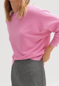 Pull côtelé rose clair avec une coupe ample et un col large, accompagné d'un pantalon à carreaux noirs et blancs texturé.
