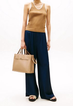 Brązowy top na cienkich ramiączkach z białą lamówką, w połączeniu z granatowymi szerokimi spodniami. Trzyma beżową torebkę z zamykaną kieszenią i górnymi uchwytami. Czarne sandały.