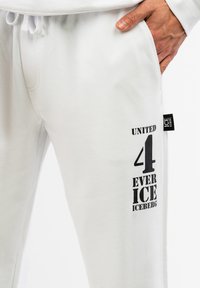 White sweatpants featuring a black printed design with "UNITE 4 EVER ICE ICEBERG" on the left leg, made of soft fabric with an elastic waistband.