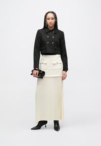 Giacca nera corta con chiusura doppiopetto a bottoni, abbinata a una gonna midi plissettata color crema e stivaletti neri alla caviglia. Tiene una piccola pochette nera.