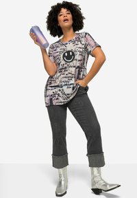 T-shirt graphique avec un visage souriant, motif en texte violet clair et noir, associé à un jean noir avec des accents cloutés et des bottes argentées.