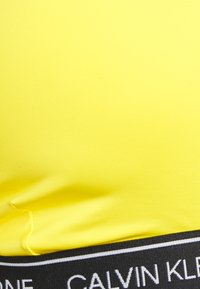 Tecido amarelo Calvin Klein com uma textura suave, apresentando uma bainha preta com letras brancas da marca e um padrão canelado.