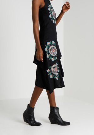 Femme portant une robe noire sans manches à plusieurs couches avec une broderie florale colorée et des bottines noires, marchant devant un fond uni.