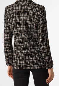 Veste en tweed noir avec un motif à carreaux beige et crème. Elle présente des manches longues et une coupe ajustée. Tissu texturé avec un col à revers.