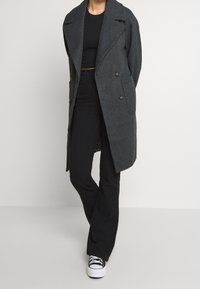 Grå ullblandad dubbelknäppt kappa med breda slag och fickor, lagd över en svart topp och svarta byxor, i kombination med vita sneakers.