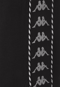 Černé tepláky s páskem po straně, na kterém jsou opakující se šedé grafické motivy sedících postav, obrysované bílými a černými pruhy.