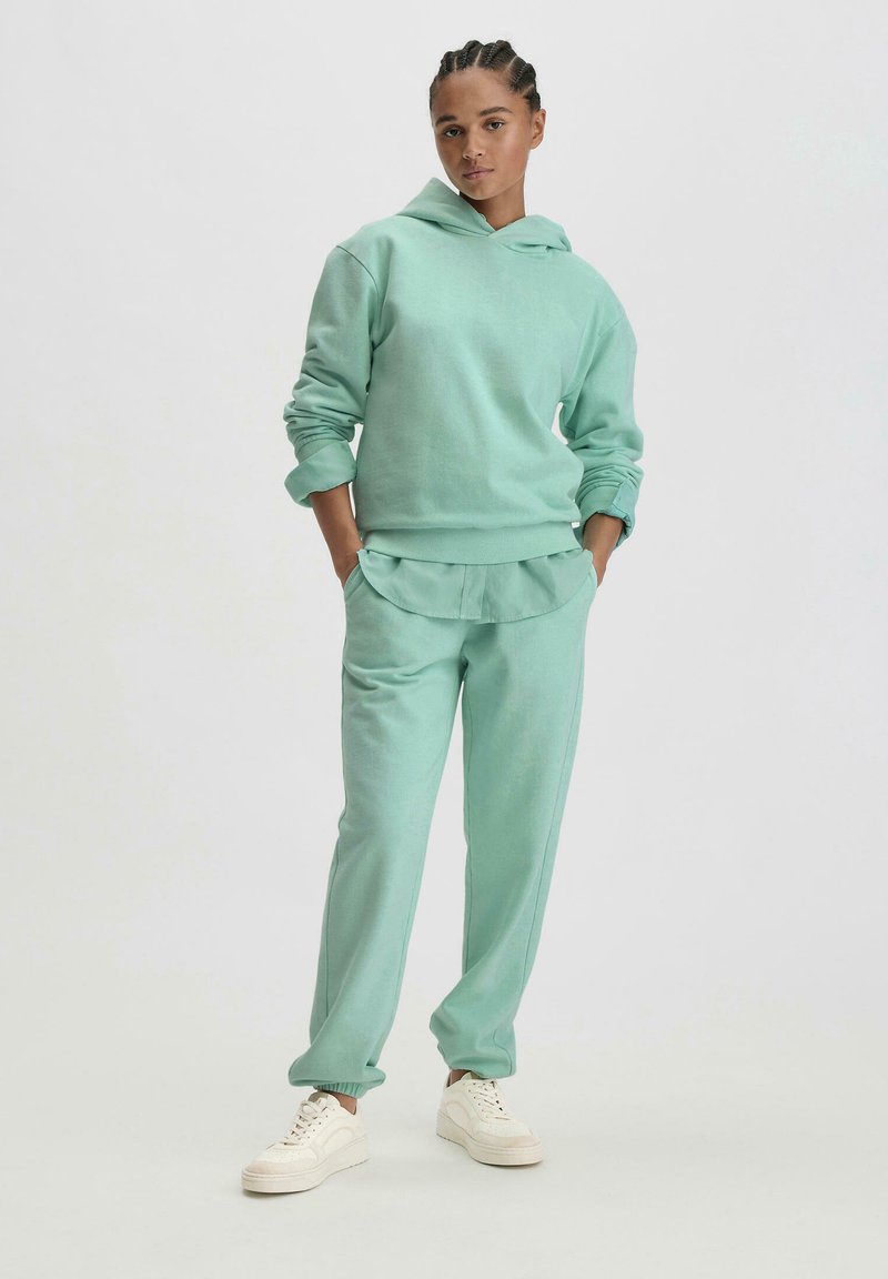 Mintgrüner Hoodie und passende Jogginghose aus weichem Stoff. Kapuzendesign mit Bündchen, Fronttaschen und lockerer Passform. Weiße Sneakers.