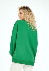 Žalias per didelis džemperis su laisvu siluetu, šonkauliniais rankogaliais ir apvadu, minkšta tekstūra ir plati iškirptė. Atgaliniai vaizdai pabrėžia paprastumą.