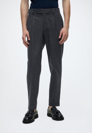 Pantalones cortos de cuadros gris oscuro para hombre combinados con mocasines negros con borlas, usados por una persona con las manos apoyadas a los lados.