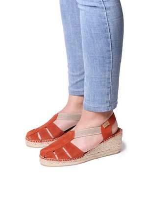 Červené semišové wedge sandály s výřezovým designem, elastickými pásky a tkanými jutovými podešvemi, předváděné na osobě v lehkých modrých džínách.
