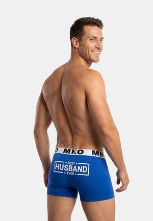 Lächelnder, oberkörperfreier Mann, der blaue Boxershorts trägt, auf deren Rückseite „Bester Ehemann aller Zeiten“ steht, schaut über seine Schulter vor einem schlichten Hintergrund.