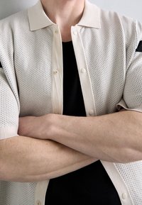 Cardigan corto color beige chiaro a maniche corte con un design a maglia texturizzata e perforata, e un colletto classico, chiuso con bottoni tondi. Maglietta nera visibile sotto.