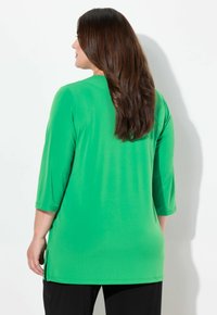 Tunika verde con maniche 3/4, realizzata in tessuto morbido. Presenta un collo rotondo e spacchi laterali. Indossata con pantaloni neri. Design semplice.