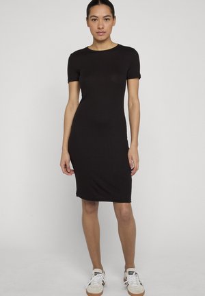 Sötét hajú nő, aki elegáns kontyba fogott hajjal, fekete, testhezálló, rövid ujjú, térd alá érő ruhát és fehér tornacipőt visel, tiszta háttér előtt áll.