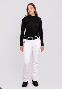 Weiße Hose mit seitlichen Reißverschlusstaschen, schwarzes Langarmoberteil und schwarzer Gürtel. Das Modell steht auf einem hellen, einfarbigen Hintergrund und trägt schwarze Schuhe.