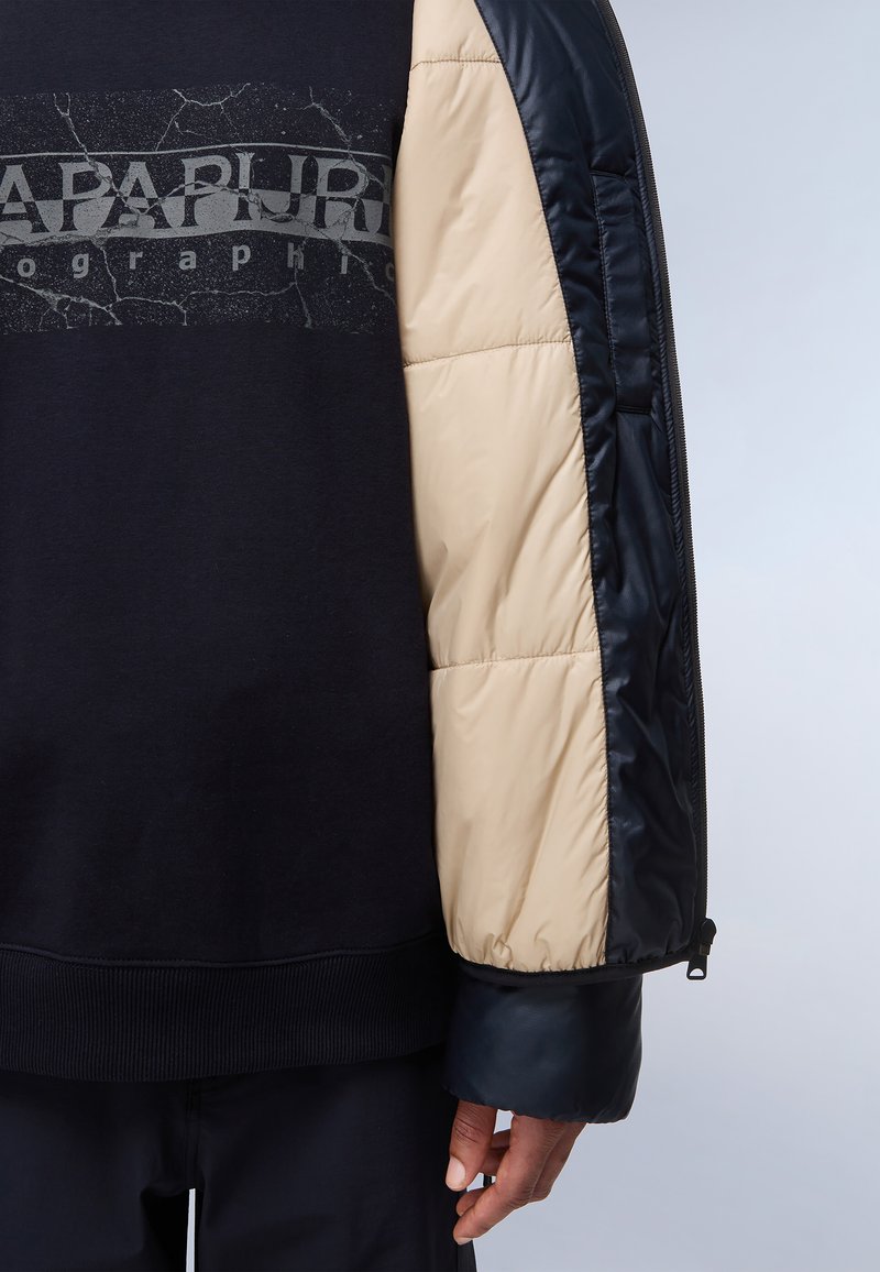 Czarny sweatshirt z szarym pękniętym logo, beżowymi i błyszczącymi niebieskimi rękawami puchowymi, ściągaczami oraz gładką teksturą. Zamek błyskawiczny na rękawie.