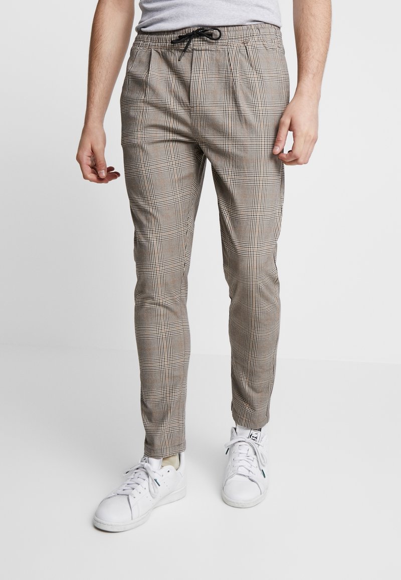 Pantalon à carreaux marron avec une coupe fuselée, taille élastique et cordon de serrage, associé à des baskets blanches. Tissu à texture lisse.