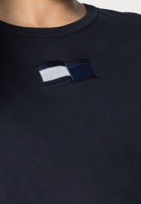 Černé bavlněné tričko s vyšitým designem dvou stylizovaných vlajek v šedé a námořnické barvě, umístěným na hrudníku.