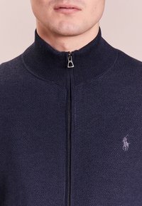 Mörkblå zip-up-tröja med strukturerat stickat mönster, hög krage och liten broderad logotyp på bröstet, med en silverfärgad dragkedja för stängning.