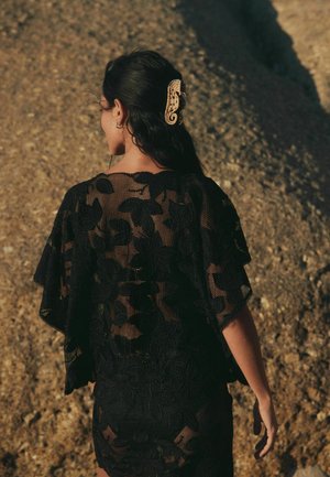 Frau mit langen dunklen Haaren, die ein schwarzes Spitzenkleid und eine haarspangenförmige Seepferdchen trägt, vor einem felsigen Hintergrund stehend.