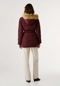Chaqueta tipo puff de color burdeos con capucha de piel sintética, cintura ceñida con cinturón, puños elásticos y diseño acolchado, combinada con pantalones de pierna ancha en crema.