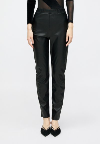Femme portant un pantalon slim en simili cuir noir, des escarpins noirs à bout pointu et un haut transparent à manches longues, se tenant devant un fond blanc.