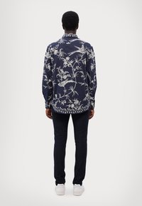 Laineen tummansininen pitkähiha-paita, jossa on valkoisia kukka- ja lintukuvioita, klassinen kaulus ja kaareva helma, yhdistettyinä tummiin farkkuihin ja valkoisiin kenkiin.