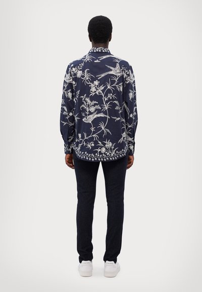 Camisa de manga larga azul marino con patrones de flores y pájaros blancos, que presenta un cuello clásico y un dobladillo curvo, combinada con jeans oscuros y zapatos blancos.