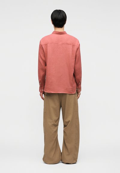 Άτομο με κοντά μαύρα μαλλιά, που φοράει ένα κοραλί μακρυμάνικο πουκάμισο και φαρδιά μπεζ παντελόνια, στέκεται γυρισμένο προς τα πίσω σε ένα απλό φόντο.