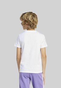 Bambino con capelli ricci biondi, vestito con una semplice maglietta bianca a maniche corte e pantaloni viola, in piedi di spalle contro uno sfondo grigio.