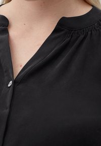 Blouse noire à boutonnage, présentant un col en V, en coton doux, avec des détails de col froncé et un unique bouton blanc près de l'encolure.