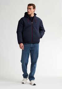 Mörkblå vadderad jacka med huva, dragkedja framtill och röda detaljer. Matchas med blå jeans och vita sneakers, stående mot en neutral bakgrund.