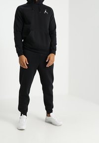 Sudadera negra con capucha y bolsillo tipo canguro, que presenta un logo blanco. Combinada con pantalones jogger negros y zapatillas blancas, muestra un estilo de ropa deportiva casual.