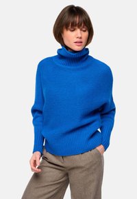 Pull en maille bleu à col roulé avec une texture côtelée et une coupe décontractée, porté avec un pantalon à motifs marron.